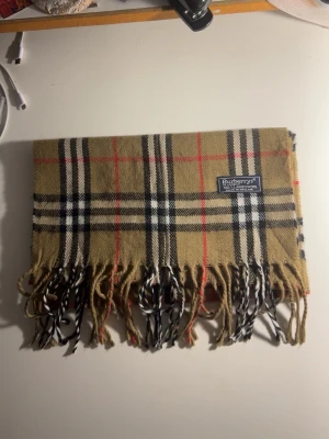 Rutig halsduk från Burberrys - Klassisk rutig halsduk från Burberrys i beige, svart, vit och röd. Tillverkad i 100% lammull och har fransar i ändarna. Den ikoniska rutiga designen gör den superenkel att styla till olika outfits.