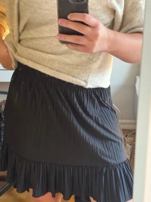 Svart kortkjol med volangkant - Säljer en svart kortkjol med plisserad struktur och volangkant längst ner. Går att vika upp om man har vill den kortare. 