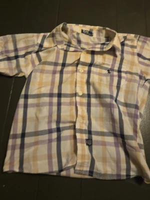 Rutig skjorta från Polo Ralph Lauren - Säljer en klassisk rutig skjorta från Polo Ralph Lauren i ljusa toner av beige, lila och marinblå. Skjortan har korta ärmar, knappar framtill och den ikoniska broderade loggan på bröstet. Perfekt för en chill och stilren look.