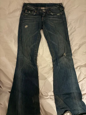 Blå bootcut jeans från True Religion - Säljer ett par blå bootcut jeans från True Religion med slitna detaljer och klassiska kontrastsömmar. Jeansen har fem fickor, låg midja och en snygg tvätt som ger en cool vintagekänsla. Perfekta för dig som gillar en avslappnad men trendig stil. innerbenslängd 75 cm