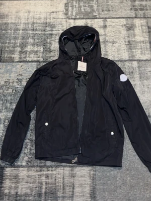 Svart vindjacka med huva från Moncler - Snygg svart vindjacka från Moncler med huva och dragkedja framtill. Jackan har två framfickor med tryckknappar och en rund Moncler-logga på ärmen. Perfekt för blåsiga dagar och enkel att matcha till olika outfits.