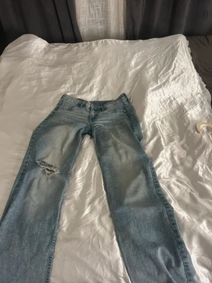Ljusblå lågmidjade jeans med slitning - Säljer ett par ljusblå jeans med flare passform och snygg slitning på vänster lår. Jeansen har klassisk femficksdesign och är tillverkade i denim med en skön känsla. Perfekta för en avslappnad och trendig look.