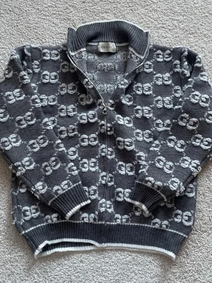 Gucci cardigan - Säljer en grå stickad kofta från Gucci med hel dragkedja och ribbad krage. Tröjan har ett upprepat GG-mönster i vitt och grått samt ribbade muddar och nederkant. Perfekt för dig som vill ha en lyxig och unik look.