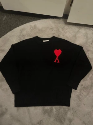Svart stickad tröja från Ami Paris  - Svart stickad tröja från Ami Alexandre Mattiussi med röd hjärtlogga och bokstaven A på bröstet. Klassisk rund halsringning och ribbade muddar vid ärmslut och nederkant. Perfekt för dig som gillar stilrena plagg med en twist.