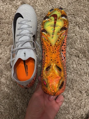 Nike Mercurial fotbollsskor vit/orange - Säljer ett par Nike Mercurial fotbollsskor med vit ovandel i stickad syntet och stora svarta swoosh-loggor. Sulan är orange med svartprickigt mönster och skimrande detaljer. Skorna har snörning och låg profil, perfekta för snabba vändningar på planen.