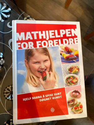 Mathjelpen for foreldre kokbok - Mathjelpen for foreldre är en norsk kokbok med hård pärm i rött och vitt. Boken innehåller recept och tips för att hjälpa föräldrar att laga hälsosam mat till barn, med färgglada bilder på olika maträtter. Perfekt för inspiration till nyttiga måltider hela dagen.