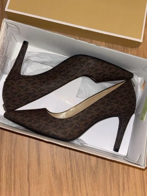 Bruna pumps med mönster från Michael Kors - Helt nya snygga bruna pumps från Michael Kors med spetsig tå och hög klack. Skorna har ett diskret MK-mönster över hela ytan och är tillverkade i syntetmaterial med en elegant finish. Perfekta för dig som vill ha en klassisk men trendig look. 