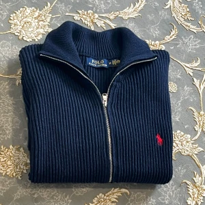 Marinblå ziptröja från Polo Ralph Lauren - Snygg marinblå stickad ziptröja från Polo Ralph Lauren med ribbad struktur och röd broderad logga på bröstet. Tröjan har hög krage, dragkedja framtill och breda muddar vid ärmslut och nederkant. Perfekt för lager på lager och stilren vardagslook.