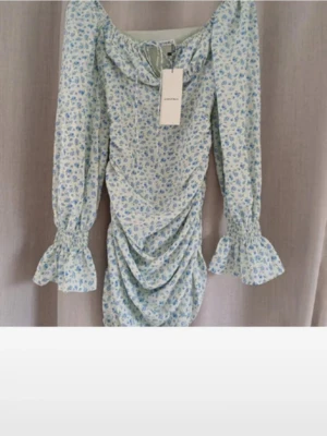 Blommig kortklänning från Adoore - Supersöt kortklänning från Adoore i ljusblått och vitt med blommigt mönster. Klänningen har långa ärmar med volang vid ärmslut, snörning i halsen och rynkade detaljer framtill. Perfekt för sommarens alla tillfällen!