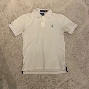 Vit piké från Polo Ralph Lauren (buda) - Klassisk vit pikétröja från Polo Ralph Lauren med korta ärmar och krage. Broderad logga på bröstet och Wimbledon-märke på ryggen. Tillverkad i bomull och har knappar framtill. Passar som en S storlek