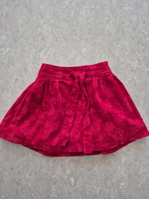 Kjol - Söt röd kortkjol med blommigt mönster och knytband i midjan. Kjolen har en lätt utsvängd form och är tillverkad i ett mjukt tyg som ger en avslappnad look. Perfekt för dig som vill ha en färgglad och lekfull stil.