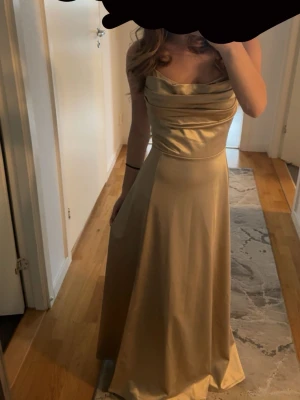 Champagnefärgad långklänning i satin - Elegant långklänning i champagnefärgad satin med smala axelband och draperad överdel. Klänningen har en figurnära passform upptill och faller mjukt från midjan. Perfekt för bal eller festliga tillfällen. Köpt utomlands bara använd en gång. Liten mini defekt (sista bild). Nypris 2499kr.