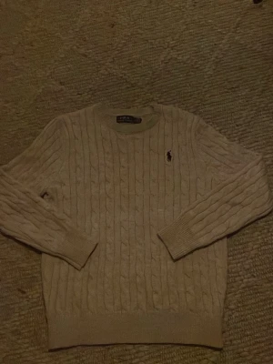 Beige kabelstickad tröja Polo Ralph Lauren - Kabelstickad beige tröja från Polo Ralph Lauren med klassisk svart broderad logga på bröstet. Tröjan har rund halsringning, långa ärmar och ribbade muddar. Perfekt för dig som gillar tidlös och clean stil. Nyskick 👍