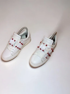 Valentino Garvani Rockstud vita sneakers - Säljer ett par vita Valentino Garavani Rockstud sneakers i skinn med röda nitar och coola detaljer på sidorna. Klassisk låg modell med snörning och rund tå. Snygga att styla till jeans eller kjol för en edgy vibe.