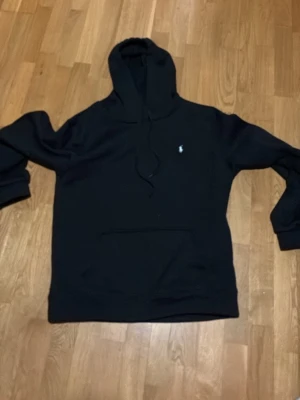 Svart hoodie från Polo Ralph Lauren - Klassisk svart hoodie från Polo Ralph Lauren med vit broderad logga på bröstet. Hoodien har huva med dragsko, stor magficka och ribbade muddar vid ärmar och nederkant. Perfekt för en avslappnad och stilren look.