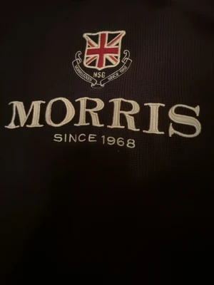 Mörkbrun tröja från Morris - Snygg mörkbrun tröja från Morris med broderad logga och brittisk flagga på bröstet. Tröjan har rund halsringning med ribbade detaljer och är perfekt för en stilren och klassisk look. Materialet känns mjukt och bekvämt.