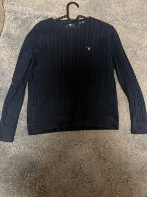Mörkblå kabelstickad tröja från Gant - Kabelstickad mörkblå tröja från Gant med rund halsringning och diskret logga broderad på bröstet. Tröjan har långa ärmar och ribbade muddar vid ärmslut och nederkant. Klassisk och tidlös design som passar till mycket. Dock ett litet hål i ärmen 
