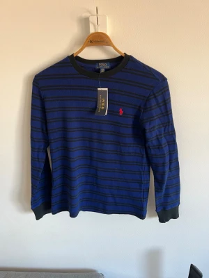 Blå randig tröja från Polo Ralph Lauren - Snygg blå långärmad tröja från Polo Ralph Lauren med tunna svarta ränder och klassisk röd logga broderad på bröstet. Tröjan har ribbade muddar vid ärmslut och halsringning. Perfekt för en stilren och avslappnad look.