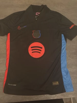 FC Barcelona svart matchtröja Nike - Snygg svart FC Barcelona matchtröja från Nike med röda och blå detaljer på sidorna. Stora Spotify-loggan på bröstet och UNHCR-tryck på ryggen. Tillverkad i Dri-FIT material som andas, perfekt för fotboll eller träning.