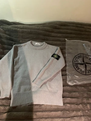 Ljusgrå stickad tröja Stone Island - Snygg ljusgrå stickad tröja från Stone Island med klassisk rund halsringning och ikonisk svart patch med gul logga på ärmen. Tröjan har ribbade muddar vid ärmslut och nederkant, perfekt för en clean och stilren look. Hör av er vid minsta lilla funderingar och pris kan alltid diskuteras!