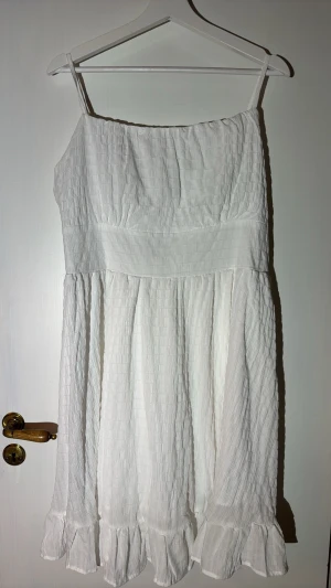 Vit sommarklänning med volang - Söt vit klänning med smala axelband och volangkant nertill. Tillverkad i ett lätt, strukturerat tyg som ger en luftig känsla. Perfekt för varma dagar och enkel att styla med accessoarer.