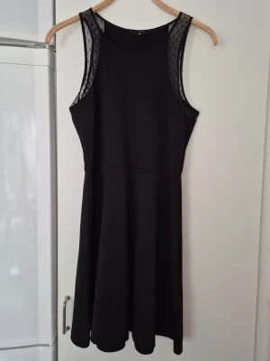 Klänning - Säljer en svart klänning från H&M i storlek M. Klänningen har ärmlös design med meshdetaljer vid axlarna och en enkel, stilren siluett. Perfekt för dig som gillar minimalistisk stil med en twist.