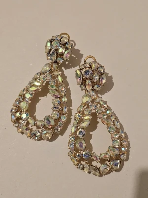 Glittriga statement örhängen i guld - Stora örhängen i guldton med massor av glittrande stenar i olika former. Örhängena har en droppformad design och är täckta av skimrande stenar som reflekterar ljuset i regnbågens färger. Perfekta för att sticka ut och ge en lyxig känsla till din look.