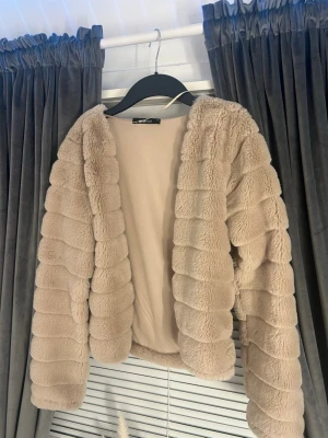 Beige pälsjacka från Gina Tricot - Säljer en fluffig beige pälsjacka från Gina Tricot. Jackan har lång ärm, öppen framtill och ett ribbat, vågigt mönster i det mjuka pälsmaterialet. Perfekt för dig som vill ha en mysig och trendig look.