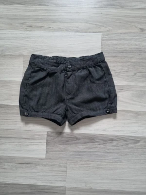 Mörkgrå shorts från Kids by Lindex - Snygga mörkgrå shorts från Kids by Lindex med hög midja och dekorativa knappar framtill och vid benslut. Tillverkade i ett mjukt tyg med diskret fiskbensmönster. Perfekta för sommarens alla äventyr!