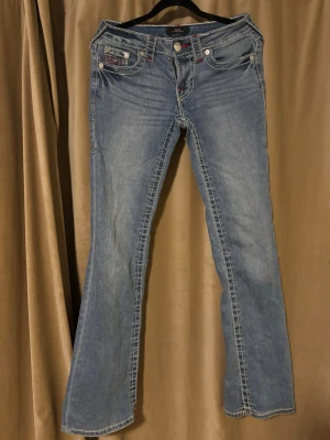 True Religion blå bootcut jeans - Säljer ett par klassiska blå jeans från True Religion med kontrastsömmar i vitt och rött. Modellen har bootcut-ben, låg midja och detaljerade bakfickor med broderad logga. Jeansen har fem fickor och är tillverkade i slitstarkt bomullsmaterial.