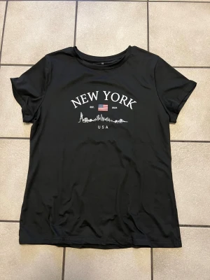 Svart New York t-shirt XL - Snygg svart t-shirt med tryck av texten 'NEW YORK', amerikanska flaggan och en linjeteckning av stadens skyline. T-shirten har rund hals och korta ärmar. Perfekt för dig som gillar streetstyle och vill ha en enkel men cool look.
