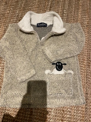 Beige stickad tröja Ramblers fårmotiv - Mysig beige stickad tröja från Ramblers med halv dragkedja och stor krage i teddyfoder. Tröjan har en ficka framtill med ett gulligt fårmotiv som sticker upp. Perfekt för dig som gillar unika detaljer och mjuka material.