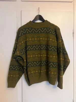 Grön vintage stickad tröja mönstrad - Unik stickad tröja i grönt med gult och mörkgrönt mönster. Tröjan har rund hals, ribbade muddar och ett retroinspirerat geometriskt mönster över hela plagget. Perfekt för dig som gillar vintage och vill sticka ut med en cool look.