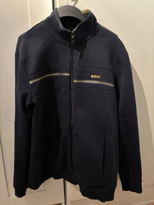Mörkblå Hugo Boss zip tröja  - Zip tröja från Hugo Boss i mjuk och följsam bomullsblandning. Modell med hel dragkedja, ståkrage och sidofickor på vardera sida. Diskreta kontrastlinjer framtill samt BOSS-logotyp på bröstet. Stilren och sportig design.  Färg: mörkblå (navy). Storlek XL (INT). Mycket bra skick, inga fläckar eller hål.  Äkta Hugo Boss – inneretiketter och detaljer finns med på bilderna.
