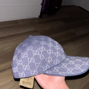 Blå GG-mönstrad keps från Gucci - Säljer en blå keps från Gucci med det klassiska GG-monogrammet över hela kepsen. Kepsen har böjd skärm och justerbar rem bak. Materialet är denimliknande tyg som ger en cool och trendig vibe. Perfekt för dig som vill sticka ut med en lyxig accessoar.