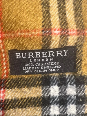 Burberry halsduk i kashmir - Klassisk halsduk från Burberry i 100% kashmir med det ikoniska rutmönstret i beige, svart, vitt och rött. Halsduken har fransar i ändarna och är tillverkad i England. Perfekt accessoar för att lyfta din stil.