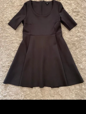 Svart kort klänning från COS - Stilren svart klänning från COS med rund halsringning och halvlånga ärmar. Klänningen har en lätt utställd kjol och dragkedja i ryggen. Tillverkad i ett slätt material som ger en snygg siluett.