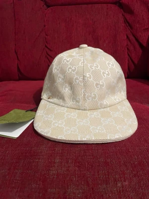 Beige keps från Gucci med GG-mönster - Snygg beige keps från Gucci med klassiskt GG-monogram i vitt över hela kepsen. Kepsen har böjd skärm och är tillverkad i canvasmaterial. Perfekt accessoar för att lyfta din outfit med en lyxig touch.