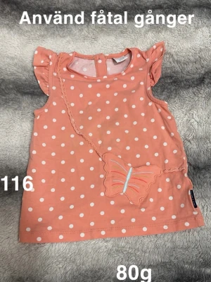 Korall/apricosfärgad prickig topp med fjärilsdetalj - Supersöt korall/apricosfärgad topp med vita prickar och en dekorativ fjäril framtill. Toppen har volangdetaljer vid axlarna och knapp i nacken. Perfekt för dig som gillar gulliga detaljer och lekfull stil.🌸 Fint använt skick. Storlek 116. 