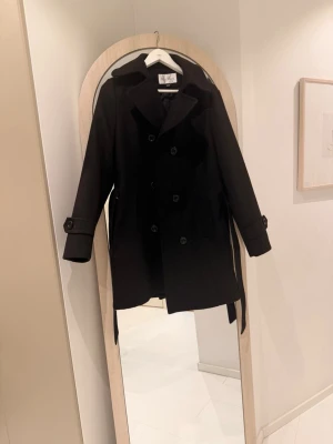 Svart kappa med bälte  -  dubbelknäppning, bred krage och två fickor framtill. Kavajen har dekorativa knappar vid ärmsluten och ett bälte i midjan. Klassisk modell i ullblandning som passar perfekt till höst och vinter.