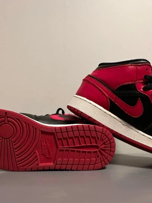 Air Jordan 1 Mid Svart/Röd - Snygga Air Jordan 1 Mid sneakers i svart och rött läder med klassisk röd Swoosh och svart snörning. Skon har vit mellansula och röd yttersula med ikoniskt mönster. Perfekt för dig som gillar streetwear och vill sticka ut med en tidlös modell.