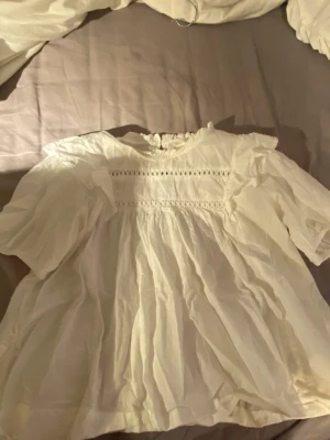 Vit romantisk blus med spetsdetaljer - Söt vit blus med korta puffärmar och fina spetsdetaljer framtill. Blusen har en liten volang vid halsen och knappstängning i nacken. Perfekt för en romantisk och feminin look. Tillverkad i ett lätt och luftigt tyg.