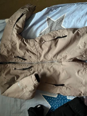 Beige täckbyxa med dragkedjor - Beiga täckbyxor i overallmodell med flera svarta dragkedjor och huva. Byxorna har förstärkta knän och resår vid ärm- och benslut. Perfekta för kalla dagar och utomhusaktiviteter. Materialet är slitstarkt och vattenavvisande.