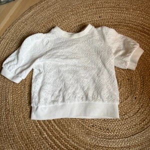 Vit topp från Holly & Whyte - Vit topp från Holly & Whyte med korta ärmar och diskret strukturmönster i tyget. Modellen har breda muddar vid hals, ärmslut och nederkant. Perfekt för en clean och fräsch look.