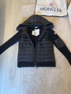  Moncler cardigan - Snygg svart dunväst från Moncler med huva och dragkedja framtill. Västen har två fickor med lock och Moncler-logga på ärmen. Materialet är en kombination av dun och stickat tyg på ärmarna, vilket ger en modern och sportig look. Jackan är helt ny!