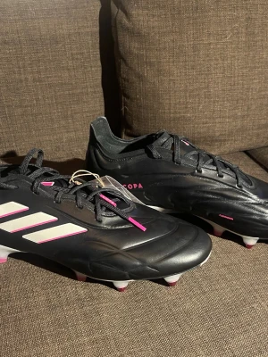 Elite Adidas Copa pure 1 SG  - Snygga svarta Adidas Copa fotbollsskor med vita och rosa detaljer. Skorna har snörning och en modern design med dobbar för optimalt grepp på planen. Perfekta för fotbollsspelare som vill ha stil och funktion. Orginal pris 2700kr. Helt nya!!! Aldrig  använda.