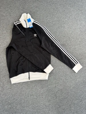 Svart Adidas track jacket med vita detaljer - Snygg svart track jacket från Adidas med klassiska vita ränder längs ärmarna och vita muddar. Jackan har dragkedja framtill, hög krage och två sidofickor. Tillverkad i polyester och har Adidas-logga på bröstet. Perfekt för en sportig och stilren look.