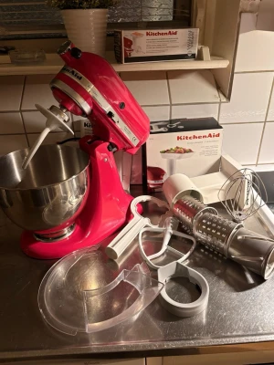 Röd KitchenAid köksmaskin med tillbehör - Säljer en röd KitchenAid köksmaskin med rostfri bunke och flera tillbehör som visp, degkrok, flatblandare, stänkskydd och grönsaksskärare. Perfekt för bakning och matlagning. Maskinen är i metall och plast och har klassisk KitchenAid-design.
