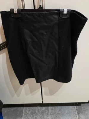 Svart minikjol med skinnimitation - Säljer en svart minikjol med panel framtill i skinnimitation och tyg på sidorna. Kjolen har en rak passform och är enkel att matcha till olika stilar. Perfekt för dig som gillar en edgy look.