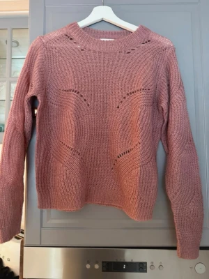 Rosa stickad tröja med hålmönster - Mysig rosa stickad tröja med rund hals och dekorativa hålmönster framtill. Tröjan har lång ärm och ribbade muddar vid ärmslut och nederkant. Perfekt för lager på lager och ger en avslappnad men trendig look.